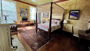 A four-poster bed with a floral bedspread, a wooden floor, a large rug, and a painting on the wall. - George Baldwin House Suite (Savannah)