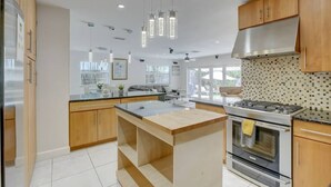 Una cocina moderna con armarios de madera, una isla central, electrodomésticos de acero inoxidable, iluminación colgante y ventanas amplias.