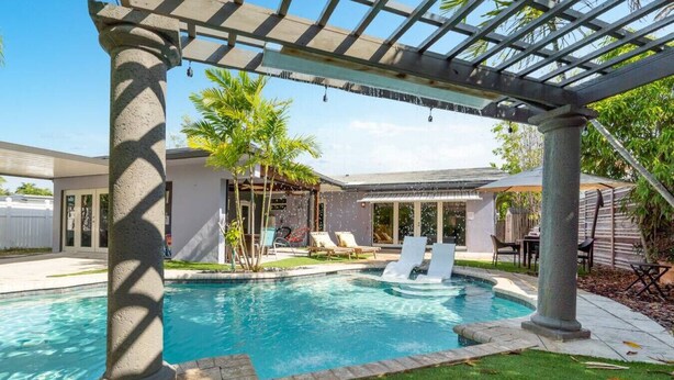 Una piscina con una pérgola, palmeras y un área de patio con sillas y una mesa.