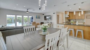 Una cocina moderna con armarios blancos, un comedor con mesa de madera y sillas blancas, y un amplio ventanal con vistas a una piscina.