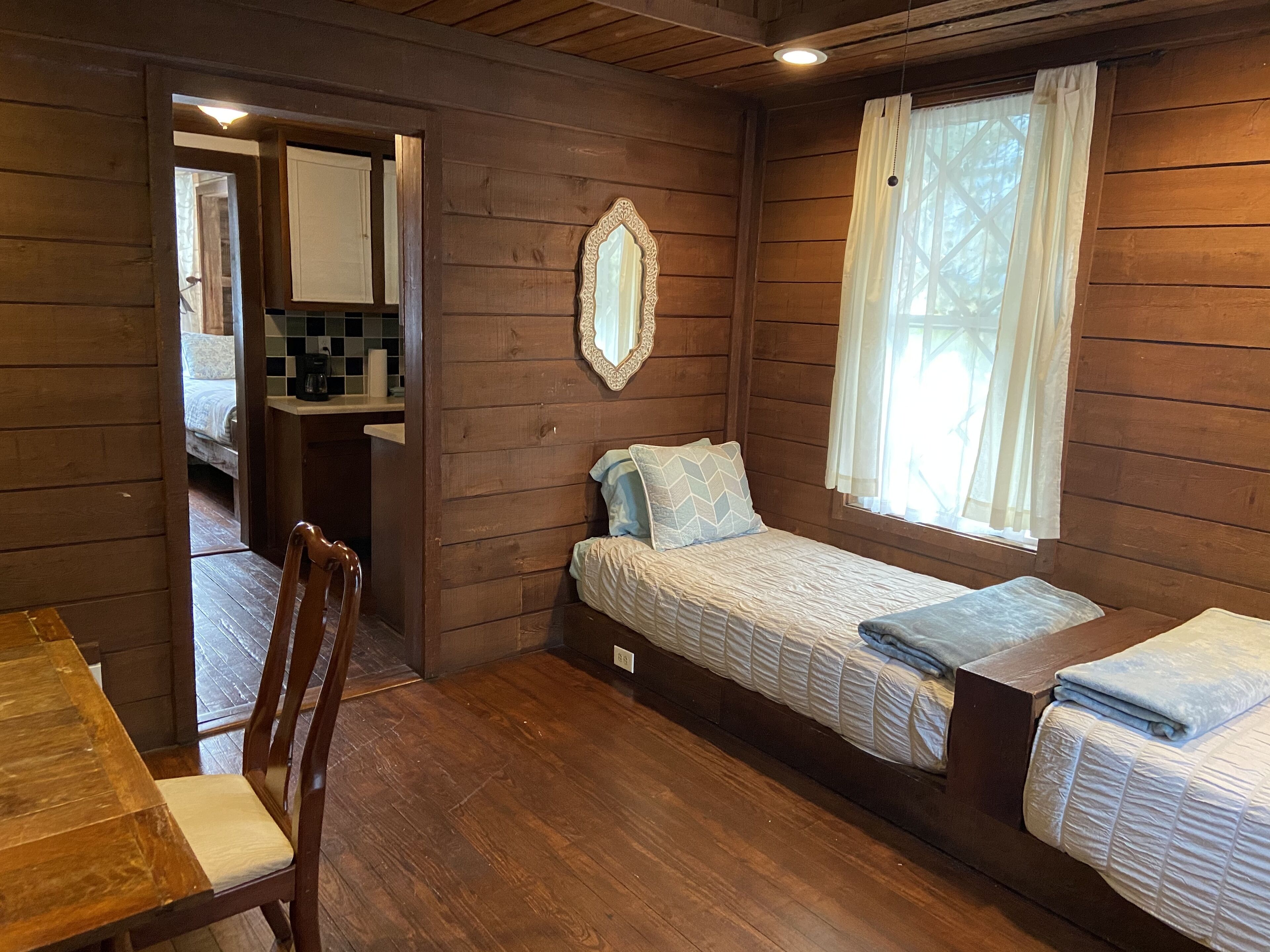 Una habitación compacta con suelo de madera, cama con colcha, mesa de madera, una silla, un espejo y una ventana con cortinas.
