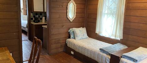 Una habitación compacta con suelo de madera, cama con colcha, mesa de madera, una silla, un espejo y una ventana con cortinas.