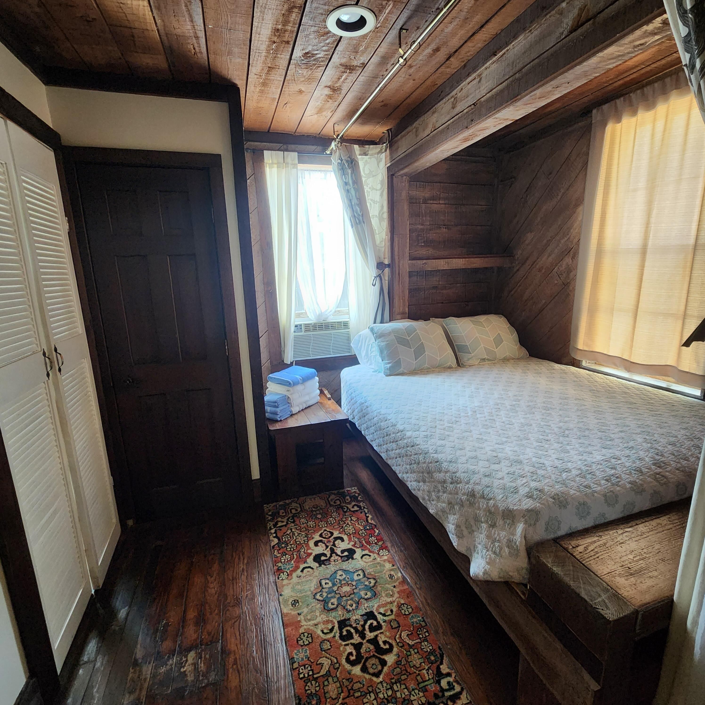 A compact room with a bed, a small table with towels, a door, and a window with curtains.
