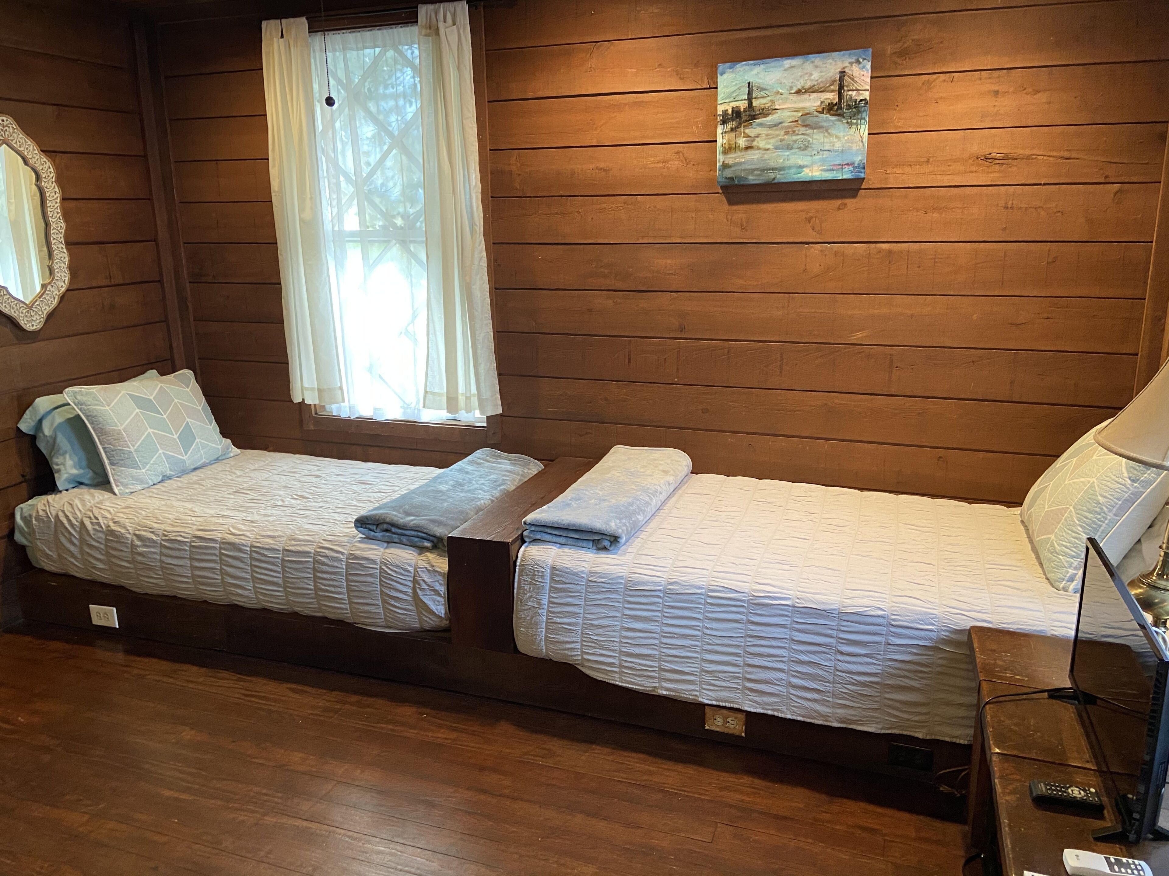 A cabin-style room with two beds, a wooden floor, a painting on the wall, and a window with curtains.