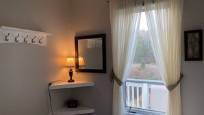 Un dormitorio con cama, lámpara de noche, espejo, ventana con cortinas y estante con ganchos.