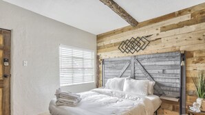 Ein Schlafzimmer mit hölzernem Kopfteil, einem Bett mit weißer Bettwäsche, einem Fenster mit Jalousien und einem kleinen Nachttisch mit Pflanze.