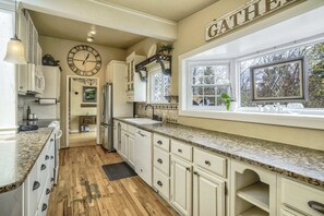 A kitchen with white cabinets, a large clock, and a window with a view of trees.