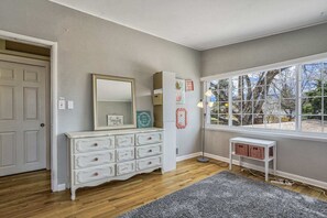 A room with a wooden floor, a white dresser with multiple drawers, a large mirror, a small table with baskets, and a window with a view of trees.