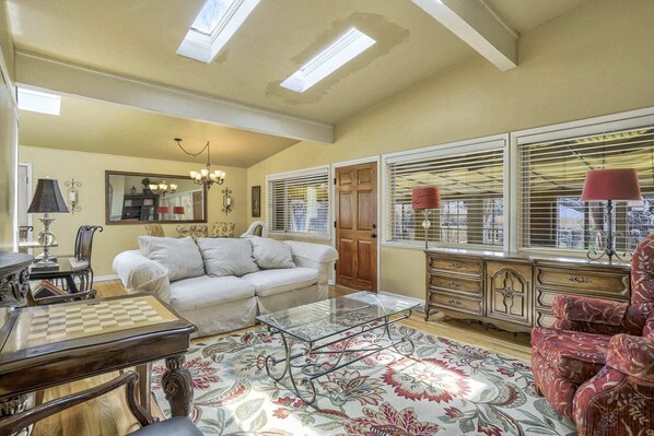 A spacious living room with a white sofa, a glass coffee table, and a red armchair. There is a wooden cabinet with drawers and a large window with blinds.