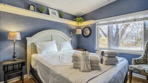 Un dormitorio con cama, mesitas de noche, lámpara, silla y ventana con vistas a árboles.