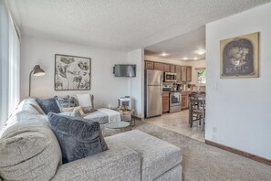 A modern living room with a sofa, a coffee table, and a TV. There is a kitchen area with a refrigerator, microwave, and cabinets.