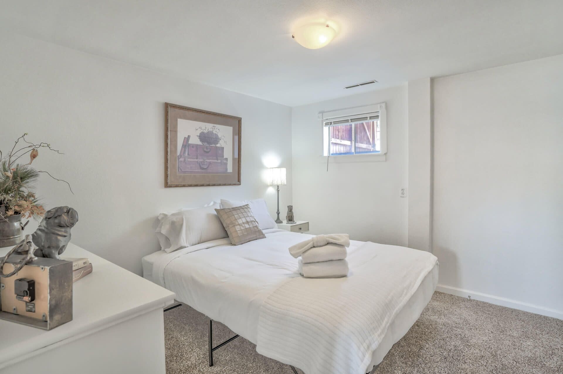 A bedroom with a bed, a bedside table, a lamp, a window, and a framed picture on the wall.