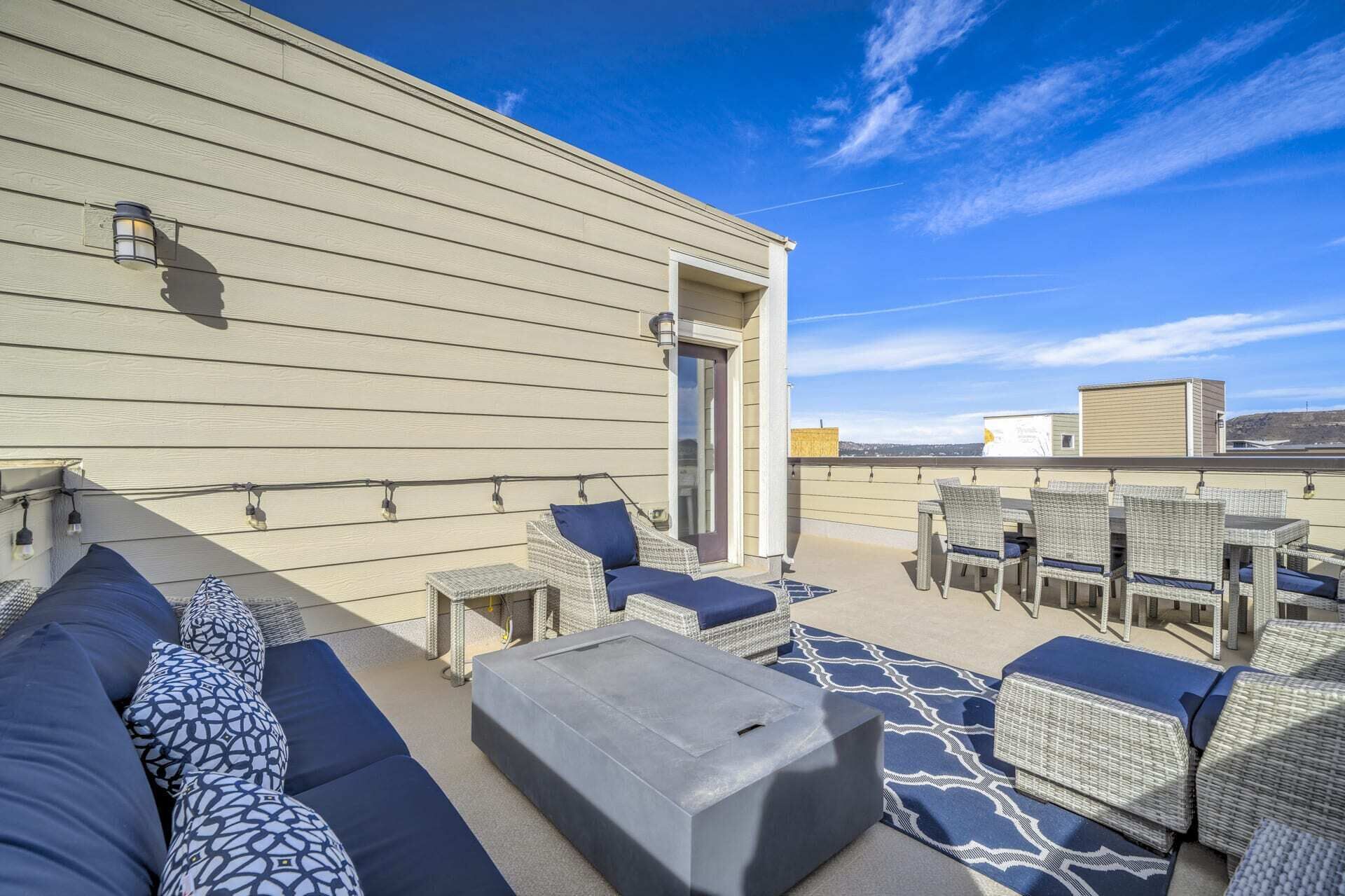 A rooftop terrace with wicker furniture, a grey ottoman, and string lights.