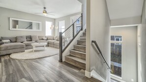 A spacious living room with a large sectional sofa, a white coffee table, and a wooden staircase with a glass railing. - 3BD Rooftop Lounge Restaurants & Parks (Castle Rock)