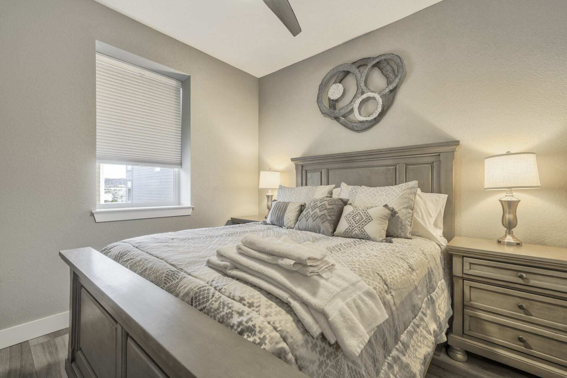 A bedroom with a bed, two bedside tables, a nightstand, a wall clock, and a window with blinds.