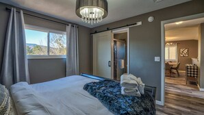 Un dormitorio con cama, un ventanal grande, una araña y un baño visible a través de una puerta abierta.
