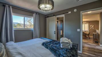 Quarto com cama, uma janela grande, um candelabro e um banheiro visível através de uma porta aberta.