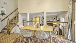 Un vaste espace dédié à la salle à manger, avec une table en bois et des chaises blanches, un escalier et une cuisine en arrière-plan.