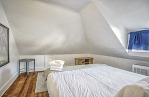 A bedroom with a sloped ceiling, a bed with white bedding, a small wooden nightstand, a window with blue curtains, and a radiator.