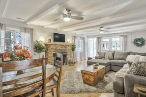 Ein geräumiges Wohnzimmer mit Kamin aus Stein, einem Holzespeisesaal mit Stühlen, einem grauen Sofa und einem Deckenventilator.