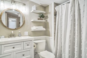 Ein Badezimmer mit weißem Waschbecken, Spiegel und Toilette. Über der Toilette befinden sich weiße Regale und ein Duschvorhang mit Muster.