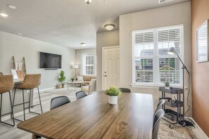 A modern living room with a wooden dining table, comfortable seating, a flat-screen TV, and a large window with blinds. - 3BD Modern Home Close to Usafa (Monument)