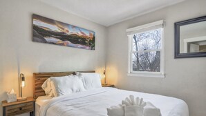Ett hotellrum med en sänggavel i trä, en stor säng med vita sängkläder, en tavla som föreställer ett landskap, ett fönster med utsikt över träd och en sänglampa.