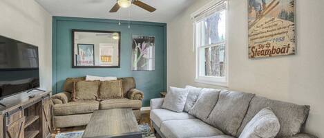 A living room with a teal accent wall, a ceiling fan, a large window, and a poster of a skier.