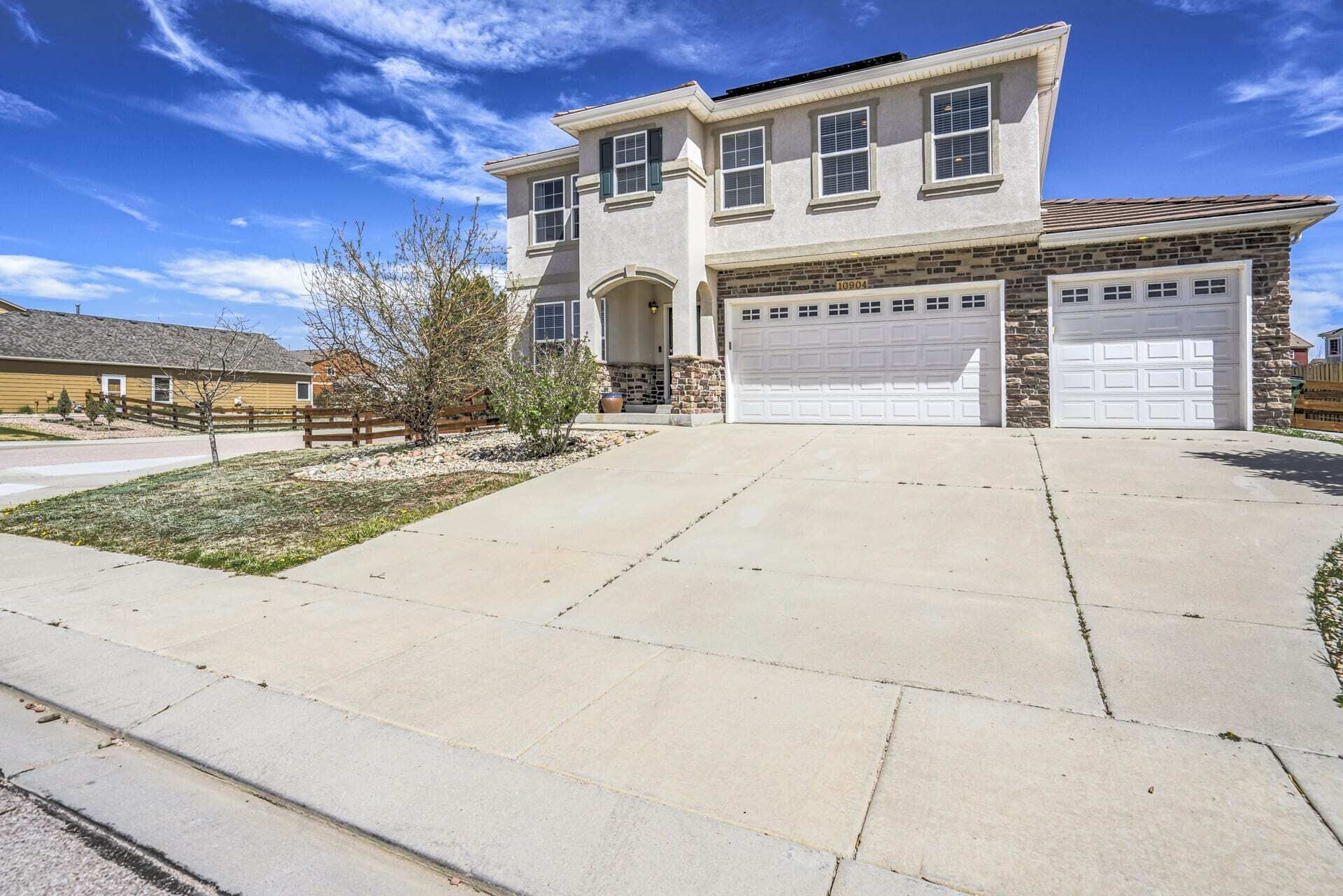 A two-story house with a stone facade, two garage doors, and a front yard with a small tree and a wooden fence.