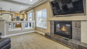 A living room with a fireplace, a TV mounted on the wall, and a kitchen area visible through an open door.