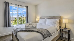 A neatly made bed with a blanket and pillows, a bedside table with a lamp, and a window with curtains.