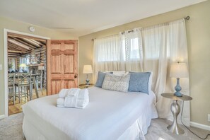 A bedroom with a large bed, two bedside tables with lamps, a wooden door, and a dining area with chairs and a table. - 3BD Cowboy Cabin Pikes Peak Mountain Log Cabin (Cascade)