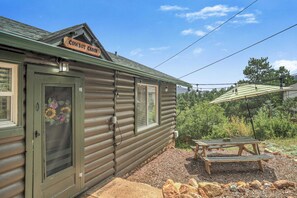A rustic log cabin with a 'Cowboy Cabin' sign, a wooden door with a floral decal, and a picnic table outside. - 3BD Cowboy Cabin Pikes Peak Mountain Log Cabin (Cascade)