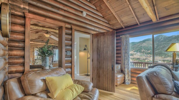 Interior de una cabaña de troncos con sofás de cuero, piso de madera y vista a un paisaje montañoso a través de un amplio ventanal.