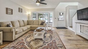 Ein Wohnzimmer mit einem beigefarbenen Ecksofa, einem Couchtisch, einem gemusterten Teppich, einem Deckenventilator und einem Fernseher auf einem Lowboard.