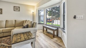 Ein Wohnzimmer mit einem beigen Sofa, einer Holzbank und einem großen Fenster mit Blick auf ein geparktes Fahrzeug draußen.