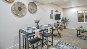 Uma sala de jantar com uma mesa posta para quatro pessoas, rodeada por cadeiras. Há cestos de vime na parede, uma TV de tela plana montada acima da mesa e um vaso de planta no canto.