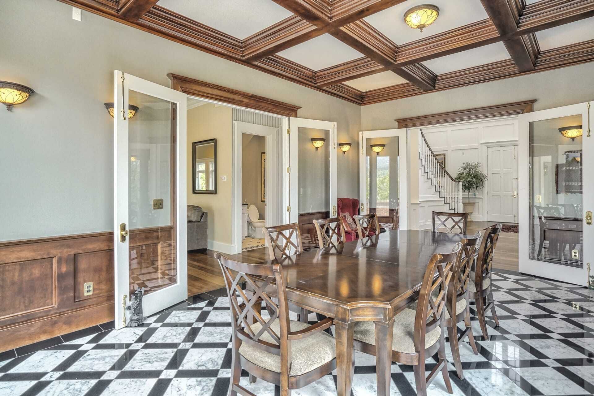 Un comedor amplio con una gran mesa de madera y sillas a juego, suelo de tablones cuadriculados y una escalera que sube a otro nivel.