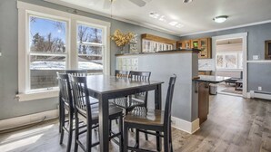 A dining area with a large table and chairs, a kitchen area with cabinets and a countertop, and large windows with a view of trees.