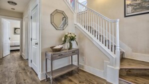 A wooden-floored hallway with a white staircase, a console table with a vase of flowers, and a framed picture on the wall.