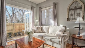 A cozy living room with a sofa, a coffee table, and a lamp. There is a window with curtains, a door, and a view of trees outside. - 1bdlovely Cottagedogs Welcome!near Olympic Training Center (Colorado Springs)