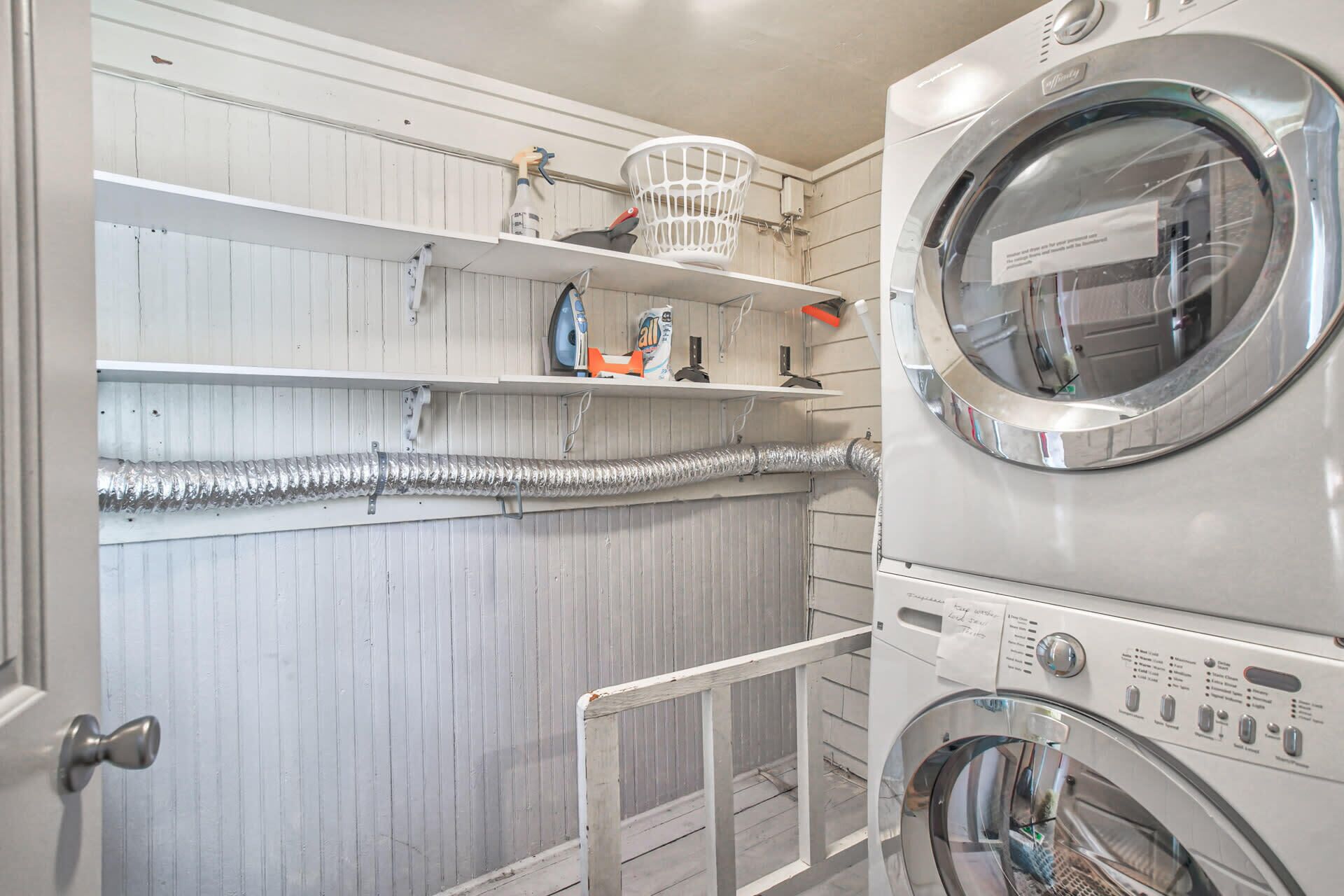 A laundry room with two stacked front-loading washing machines, shelves with cleaning supplies, and a vent running along the wall.