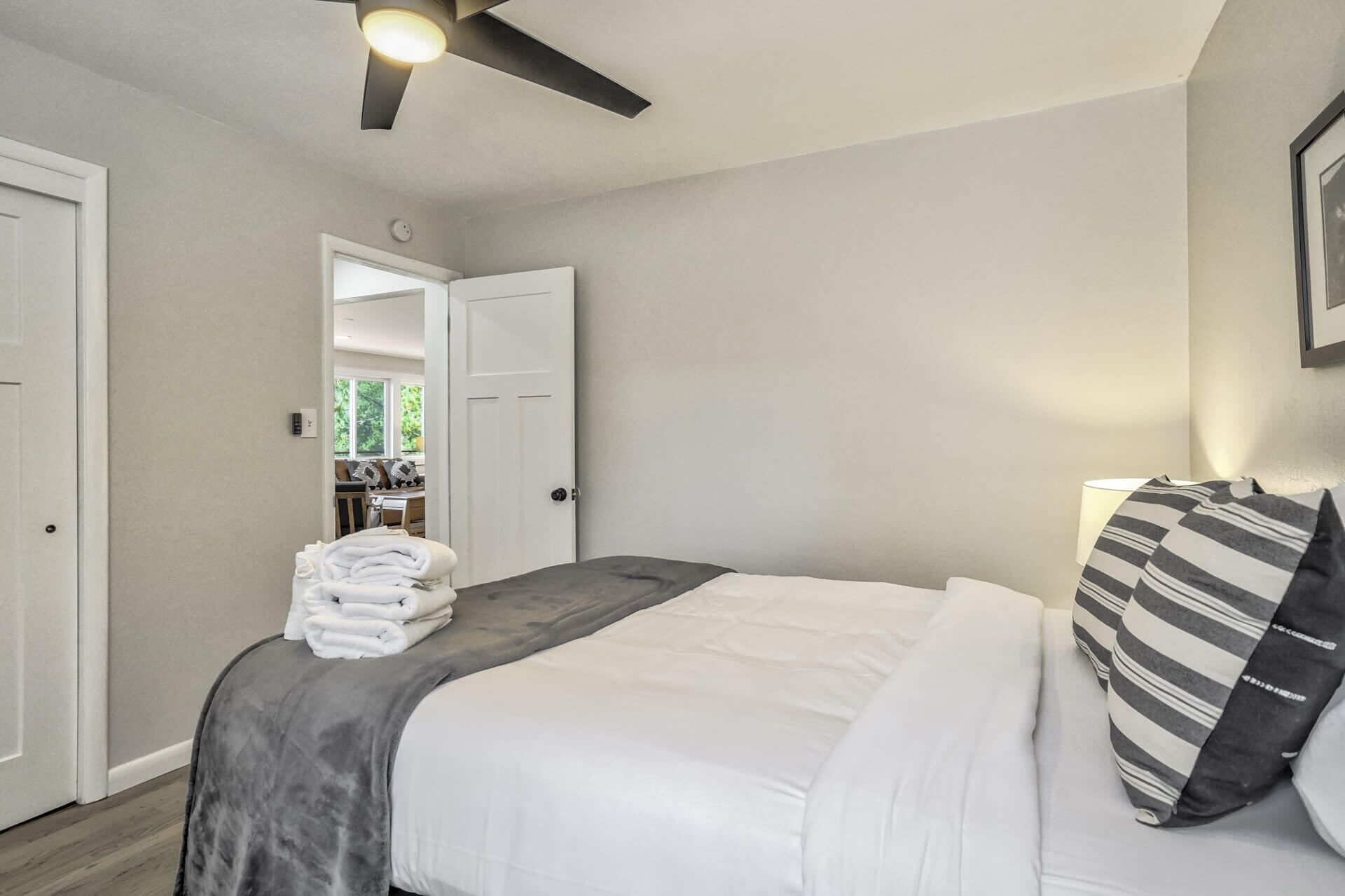 A hotel room with a bed, a ceiling fan, a bedside lamp, and a view of the adjacent room.