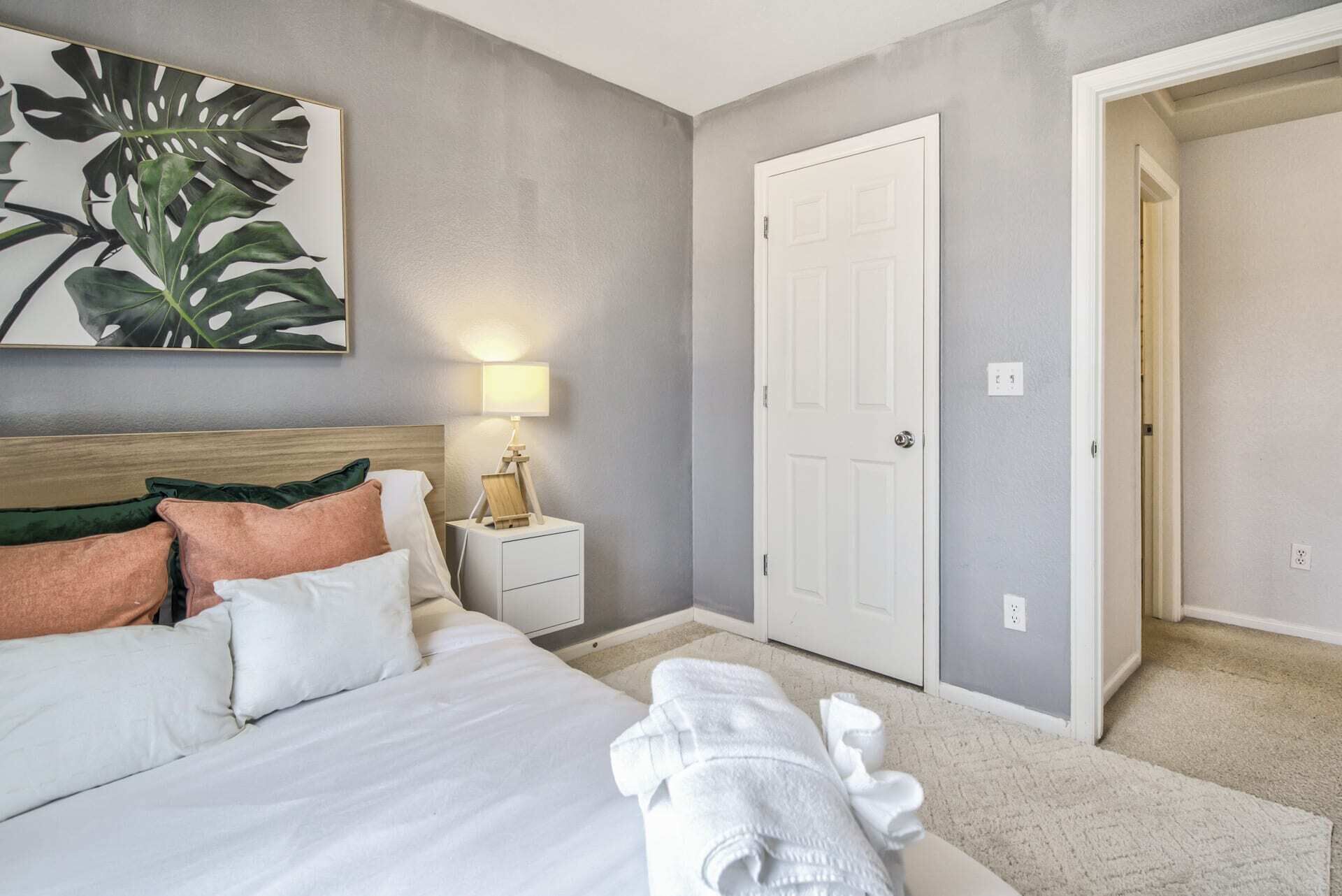 Un dormitorio con cama, almohadas, una mesita de noche con lámpara, una puerta y un pasillo.