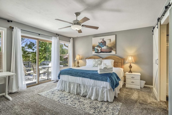 A bedroom with a bed, two bedside tables, a ceiling fan, a painting, and a view of the outdoors.