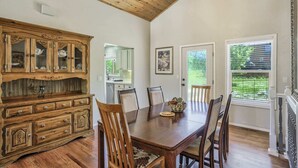 Un coin repas avec une table et des chaises en bois, un buffet en bois doté de portes en verre, et une vue sur l’extérieur par une fenêtre.