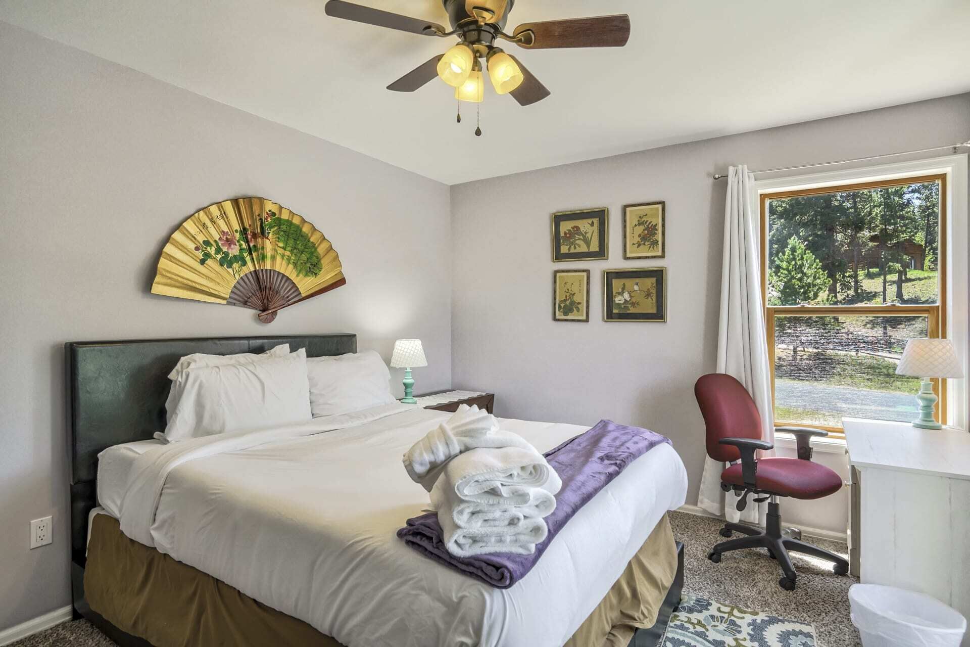 A hotel room with a bed, a desk, a chair, a ceiling fan, and a window with a view of trees.