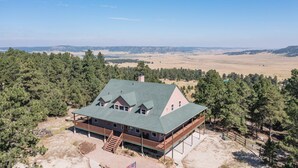 A large, two-story lodge with a green roof, surrounded by dense forest and open fields.