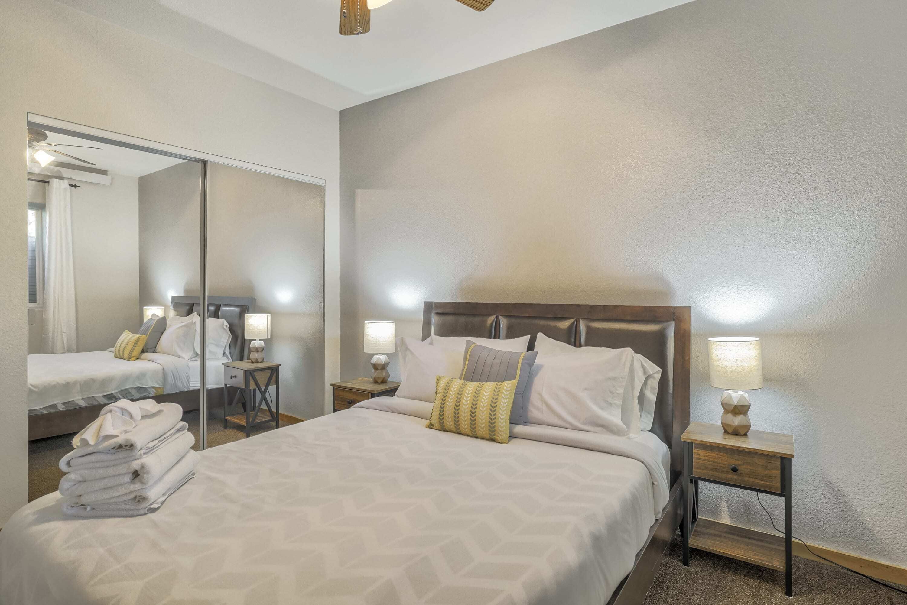 A hotel room with a large bed, a nightstand with a lamp, a mirror, and a wall-mounted shelf.