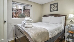 A neatly made bed with white linens, a window with a view of a rocky outdoor area, and a bedside table with a lamp and a small plant.
